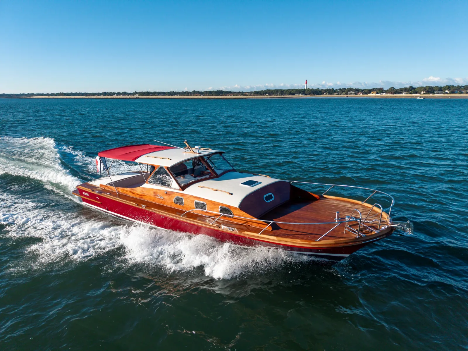 A red and orange wooden speedboat with a red canopy anchored in shallow waters off a sandy beach, with a coastal town and lighthouse visible in the background under a clear blue sky.