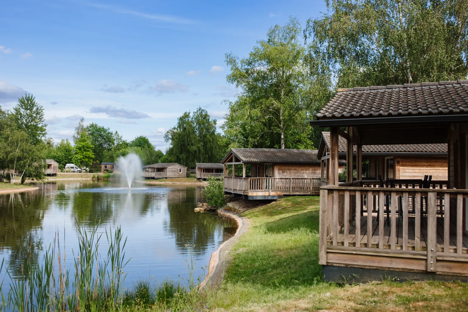Wooden holiday chalets with covered terraces overlooking a calm lake with fountain, surrounded by green grass and trees at Yelloh! Village Bordeaux Lac