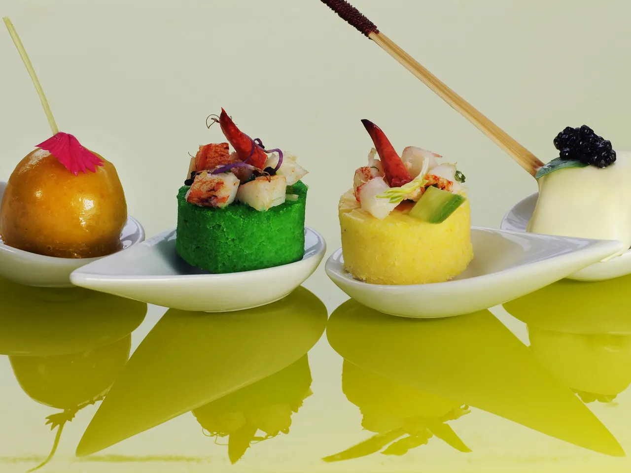 Four gourmet appetizers on white spoon-shaped plates with colored backgrounds: a golden sphere with pink flower, a green mousse with seafood, a yellow mousse with shrimp, and a white panna cotta with berries and paintbrush.
