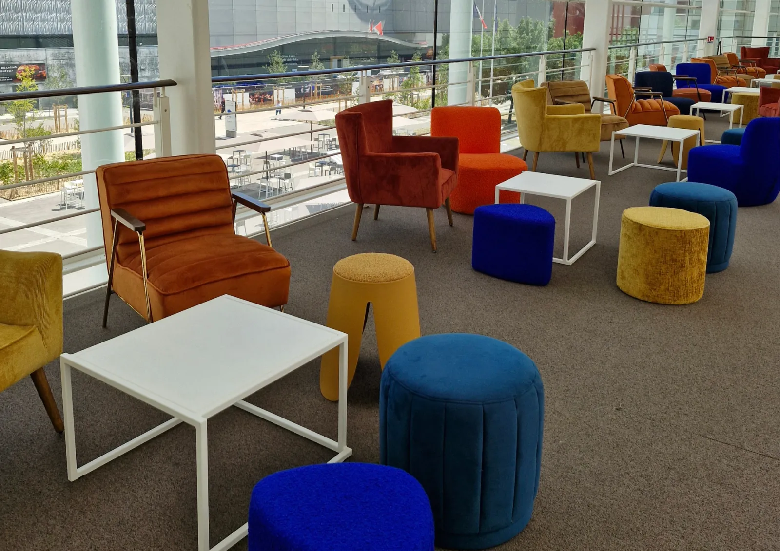 Modern lounge area with colorful upholstered furniture including orange, blue, and yellow chairs and poufs, white tables, and floor-to-ceiling windows overlooking a public plaza in Bordeaux