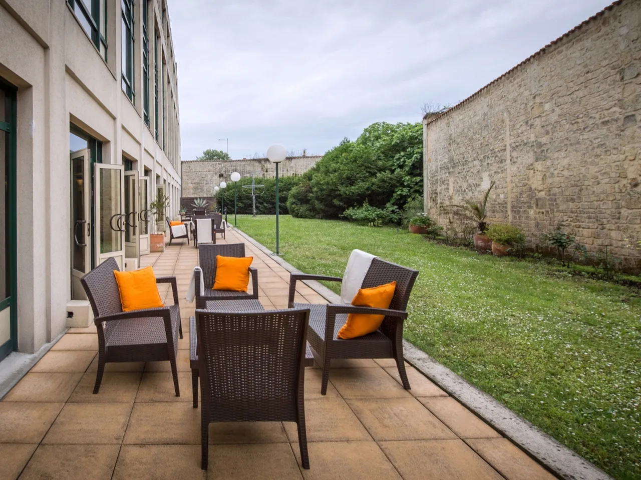 Modern hotel patio with dark wicker chairs featuring orange cushions, terracotta flooring, overlooking a manicured lawn bordered by historic stone walls at Mercure Bordeaux Château Chartrons.