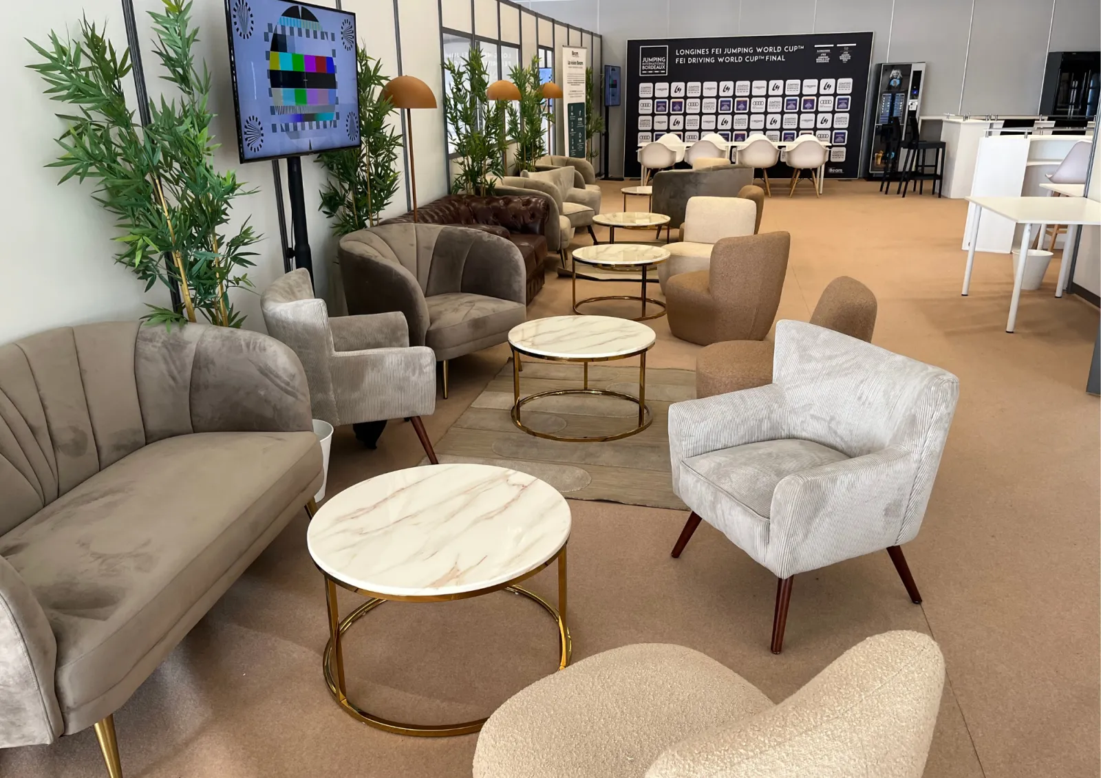 Modern lounge and showroom in Bordeaux featuring contemporary furniture displays with beige and gray upholstered chairs and sofas, marble-topped coffee tables, decorative plants, and a digital display screen showing test pattern.