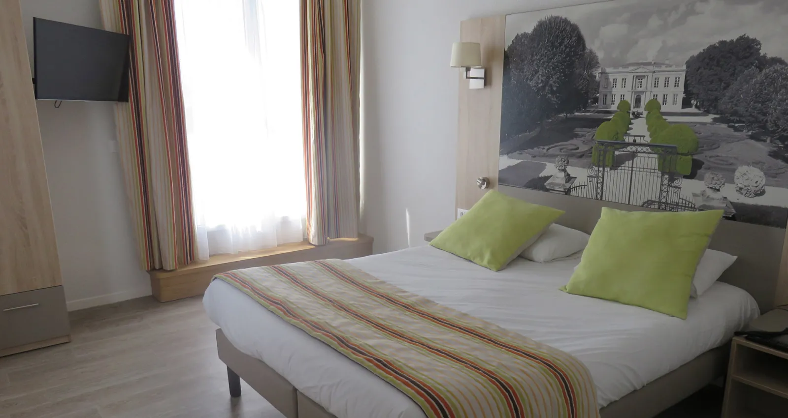 Modern hotel bedroom with a double bed featuring green cushions and striped bedding, wall-mounted TV, large windows with striped curtains, and a black and white garden photograph on the headboard wall.