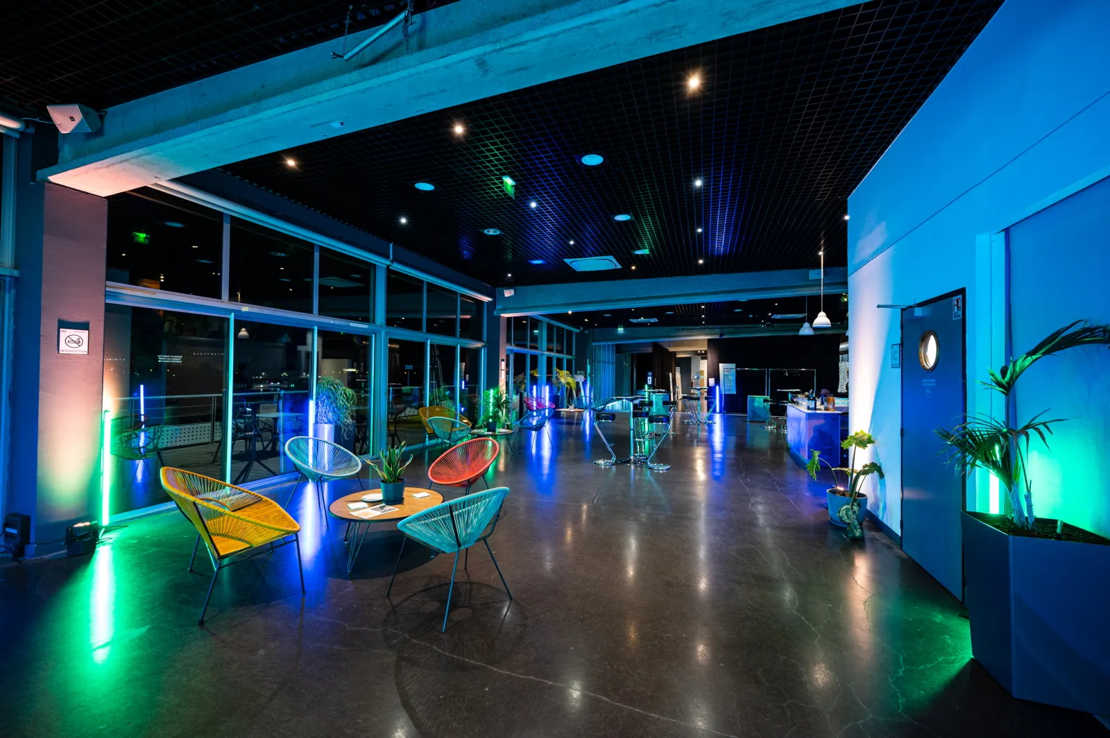 Modern lounge space at Cap Sciences Bordeaux with colorful neon lighting, contemporary seating areas, floor-to-ceiling windows overlooking the city, and green plant accents throughout.