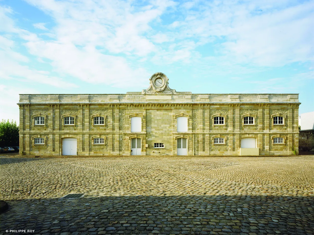 Historic stone building with clock tower and barred windows, featuring symmetrical Palladian architecture in honey-colored ashlar, located in a paved courtyard in Bordeaux