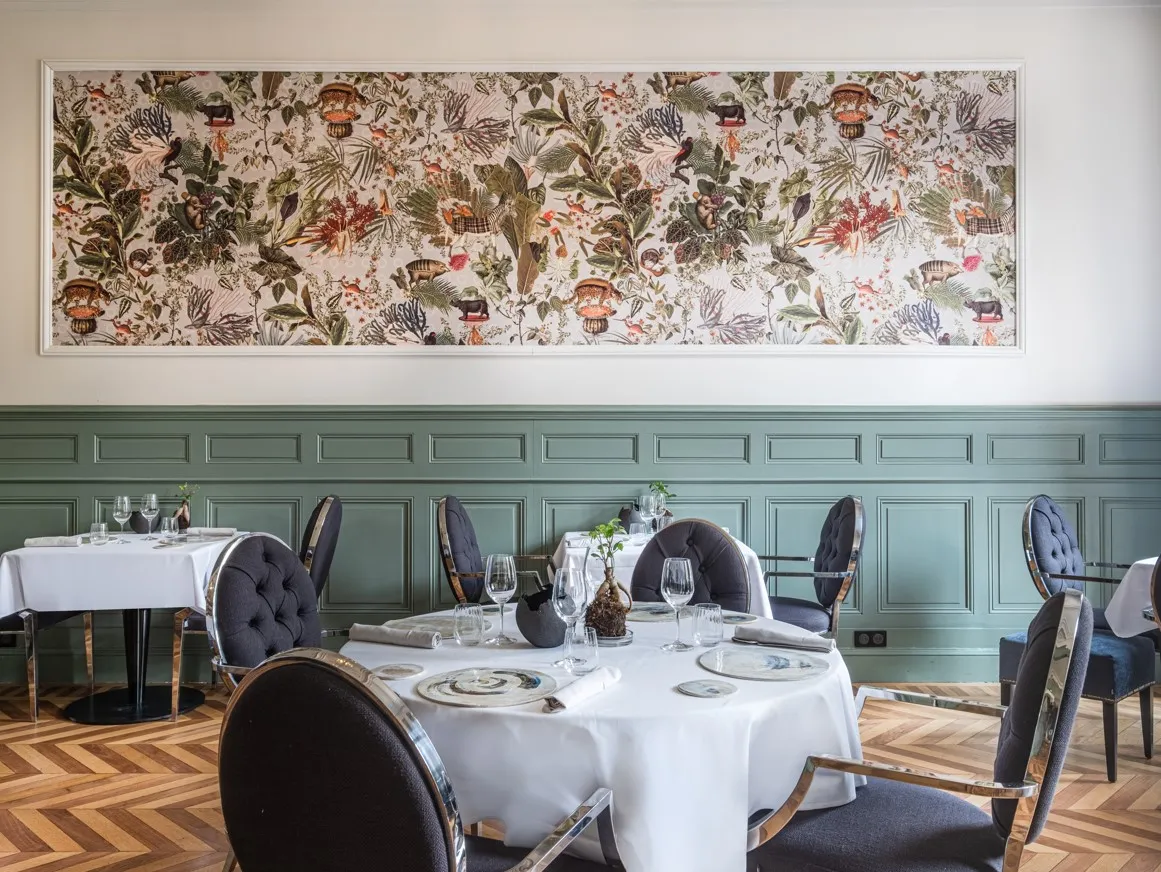 Elegant dining room at Palais Gallien Hotel & Spa in Bordeaux, featuring round tables with white tablecloths, dark upholstered chairs with wood frames, herringbone wooden flooring, sage green wainscoting, and decorative botanical wallpaper above.