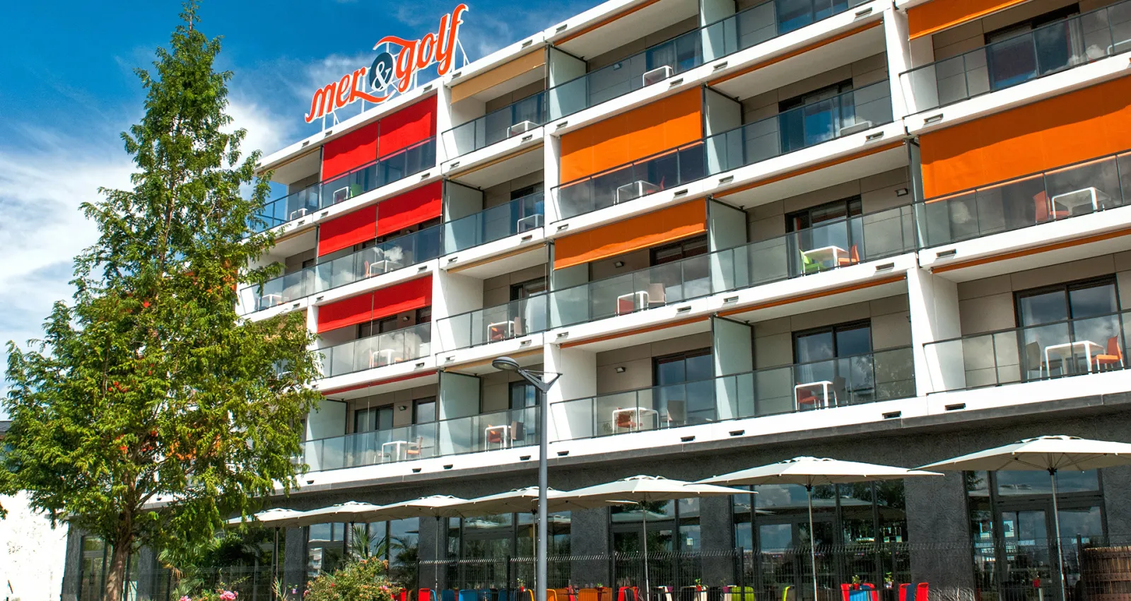 Modern apartment-hotel building with white facade, colorful orange and red balcony awnings, glass railings, and ground-floor terrace with white umbrellas and seating area. Green tree on the left side under blue sky.
