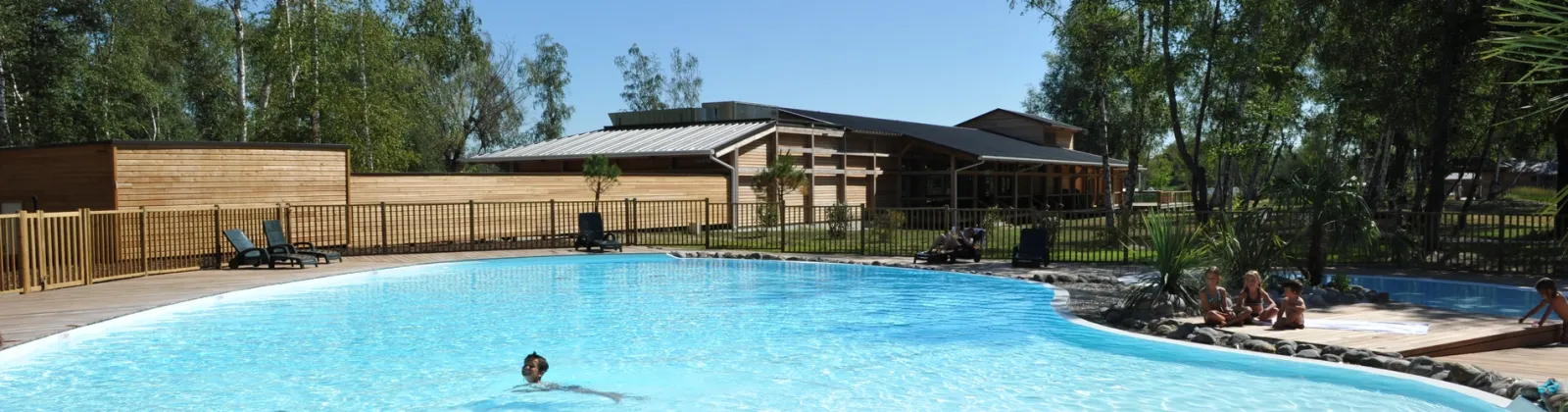 Large outdoor swimming pool with clear blue water at Yelloh! Village Bordeaux Lac, surrounded by wooden deck, lounge chairs, and landscaped grounds with trees and wooden structures in the background