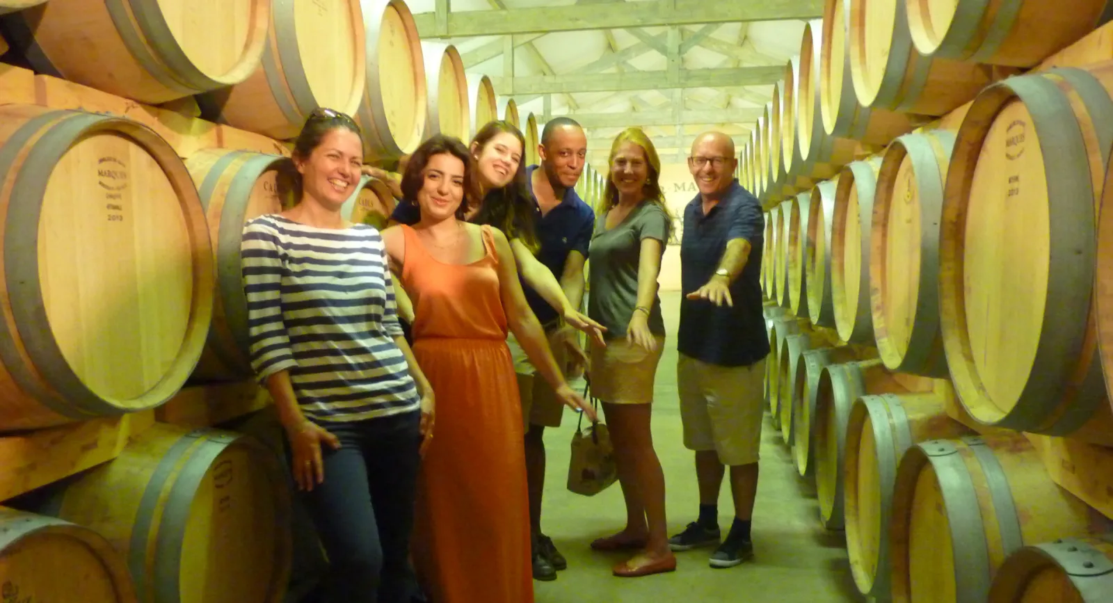 Six visitors smiling at the camera in a wine barrel cellar at Bacchus workshops in Bordeaux, surrounded by wooden oak barrels with metal bands stacked on both sides.
