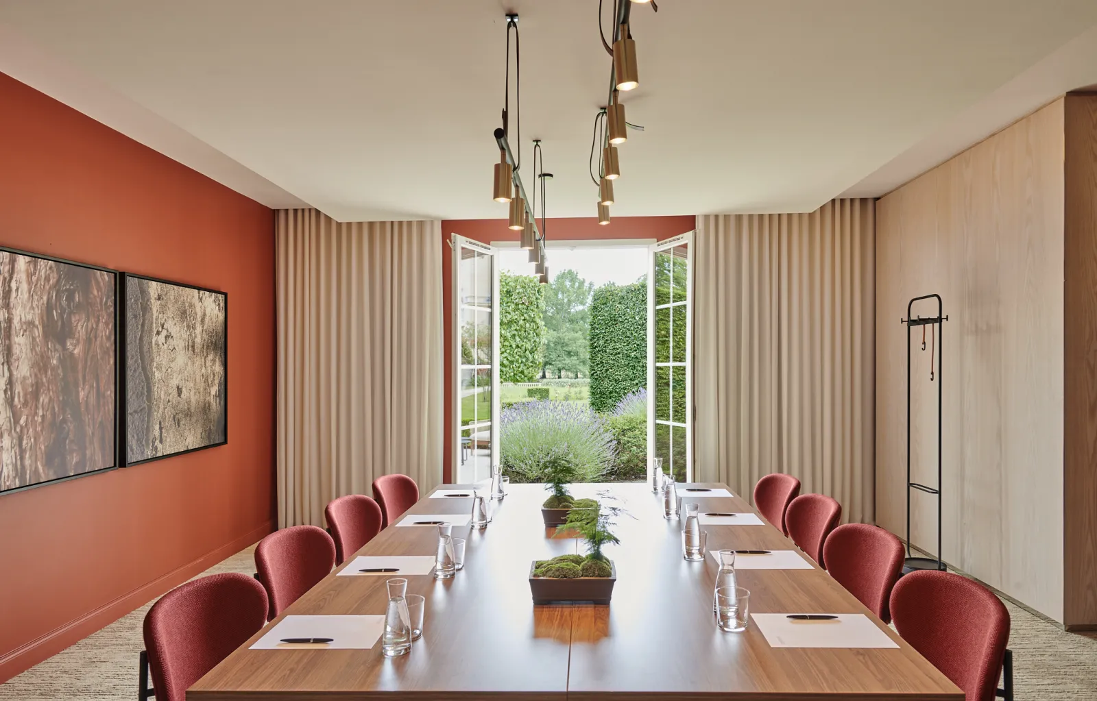 Modern conference room with long wooden table, burgundy chairs, orange accent wall, floor-to-ceiling windows overlooking gardens, pendant lights, and neutral curtains at Château Hôtel Grand Barrail in Bordeaux.