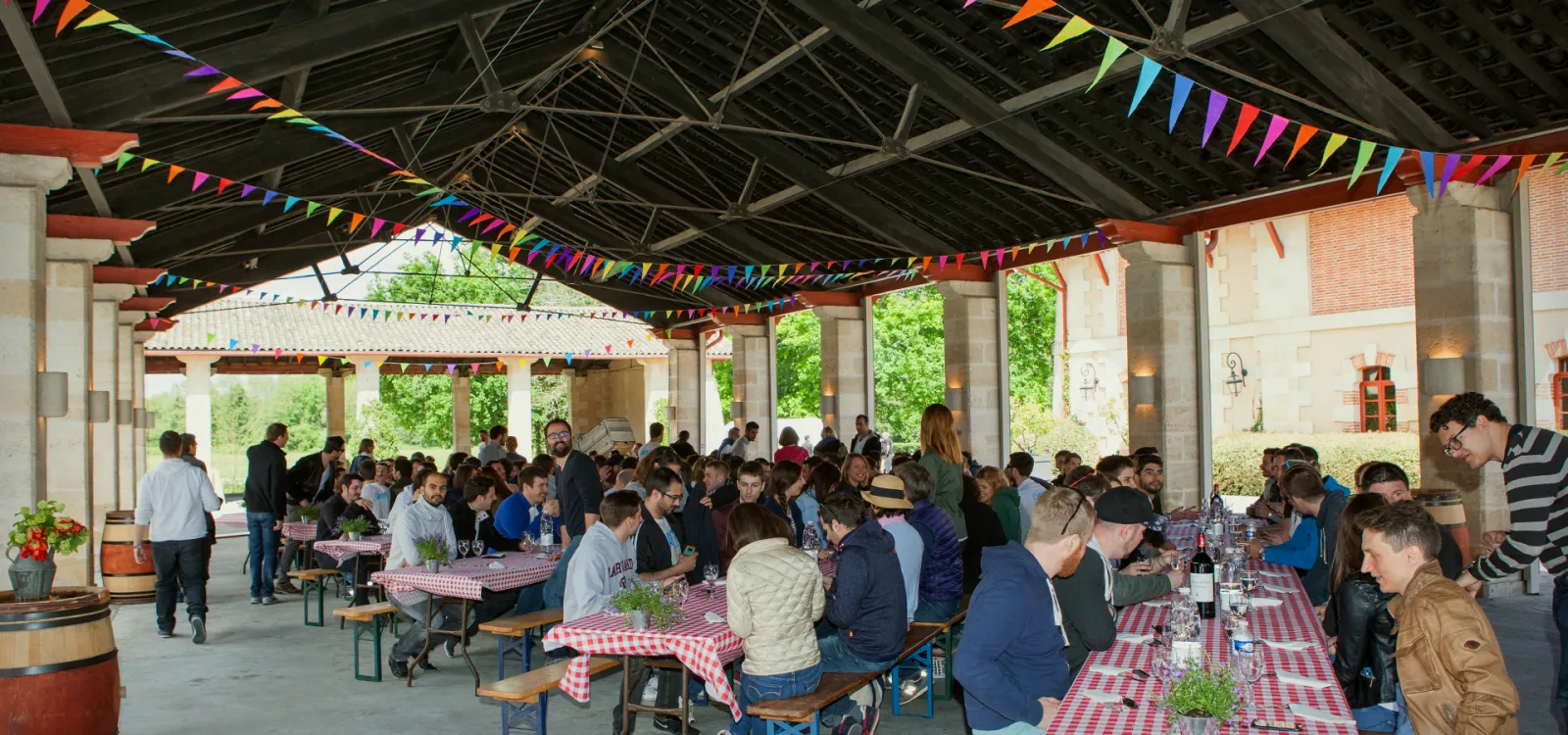 Large covered outdoor gathering space with wooden beams and colorful bunting, featuring long tables with red and white checkered tablecloths where many people are seated and socializing during what appears to be a festive community event or festival.
