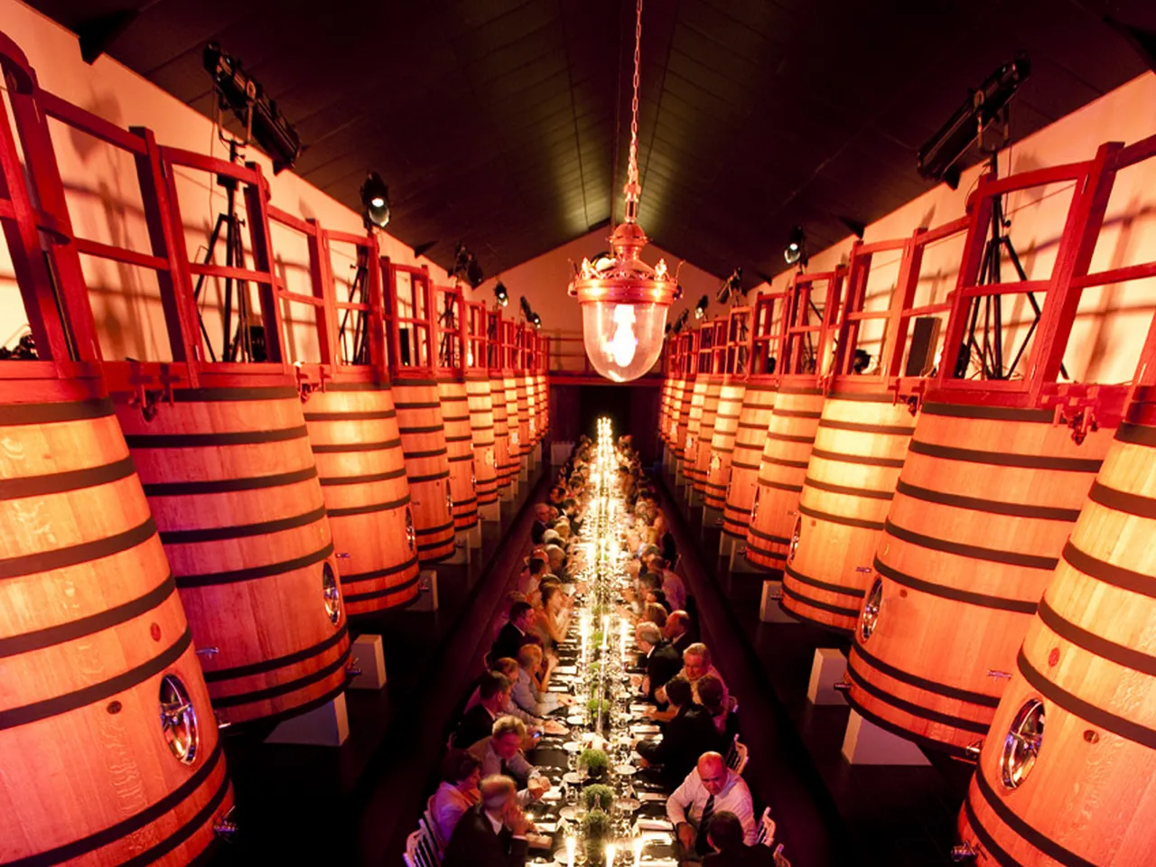 A luxurious banquet hall lined with large wooden wine barrels on both sides, with an elegant long table set for dining between them, illuminated by warm orange lighting and a central pendant lamp, at Humblot catering in Bordeaux.