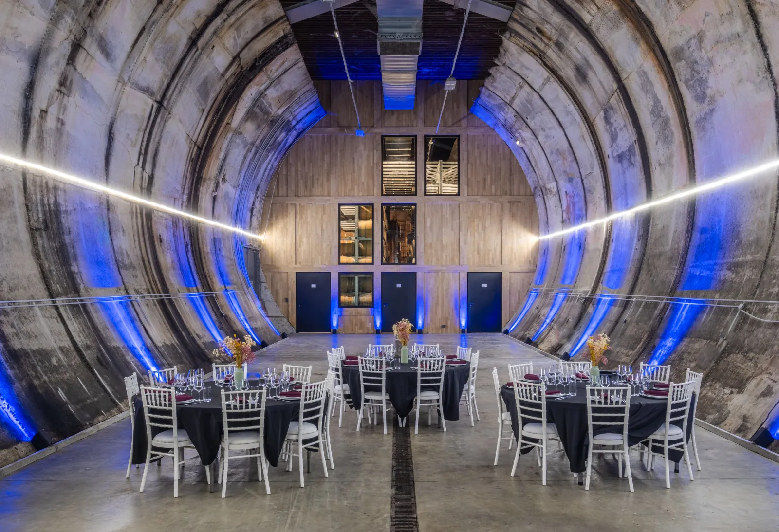 Modern event space in a converted industrial tunnel at Moon Harbour distillery in Bordeaux, featuring round tables with black tablecloths, white chairs, gold centerpieces, and blue accent lighting along the curved concrete walls.