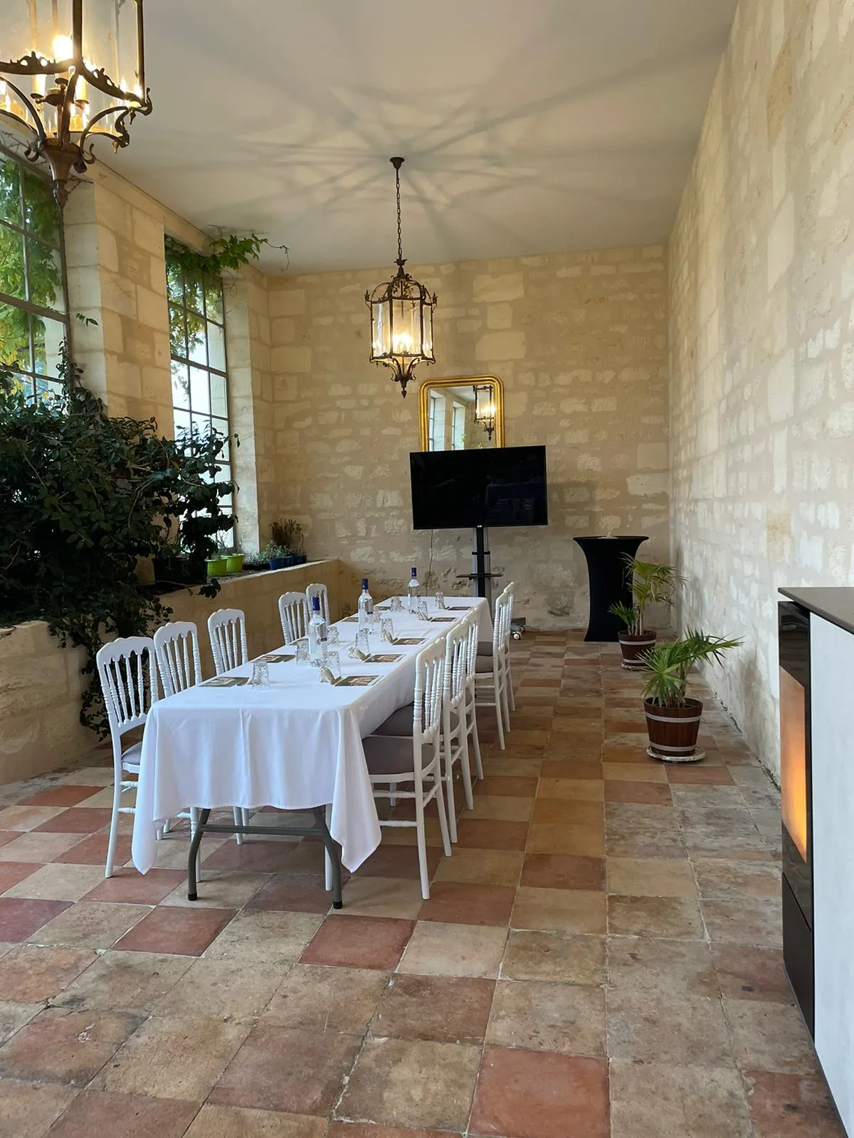 Elegant dining room at Chateau Gassies in Bordeaux with a long table set with white tablecloth and white chairs, stone walls, terracotta tile flooring, hanging lantern, and potted plants