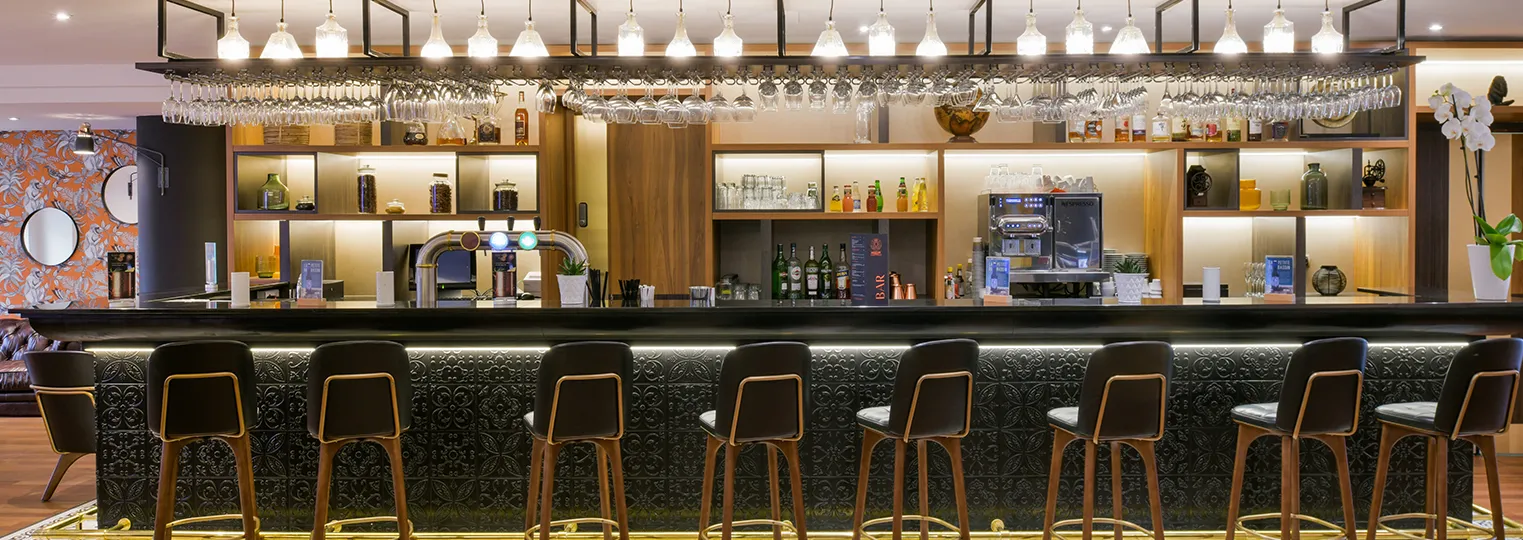 Modern hotel bar with a long black counter, wooden bar stools with gold accents, extensive shelving displaying spirits and glassware, pendant lighting, and contemporary design elements at Mercure Bordeaux Château Chartrons.