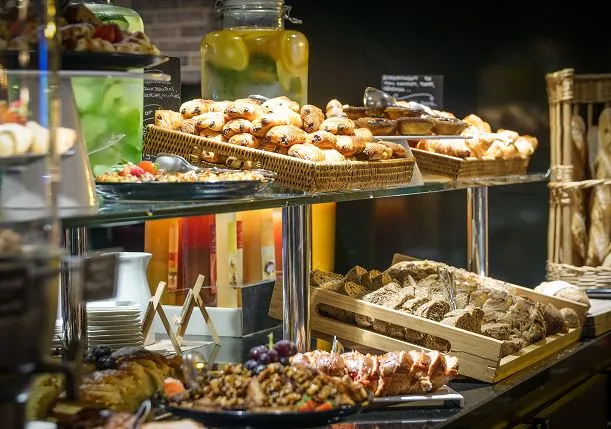 Elegant buffet display at Pullman Bordeaux Lac hotel featuring tiered glass shelves with fresh pastries in wicker baskets, bread selection, charcuterie, and beverages including infused water in glass jar