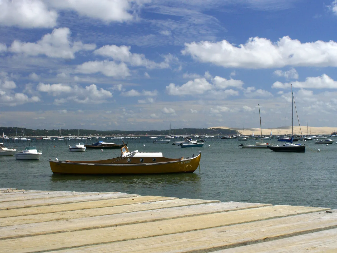 Golden rowing boat moored in turquoise waters of Arcachon Bay, with sailboats and sandy beach in foreground under blue sky with white clouds