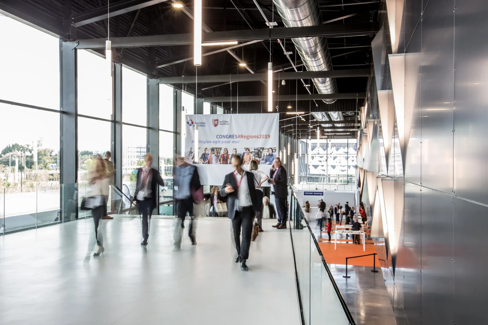 Business professionals walking through a modern conference hall at the Congres #Regions2019 event in Palais 2 l'Atlantique in Bordeaux, with floor-to-ceiling windows, contemporary lighting, and an orange reception area visible below.