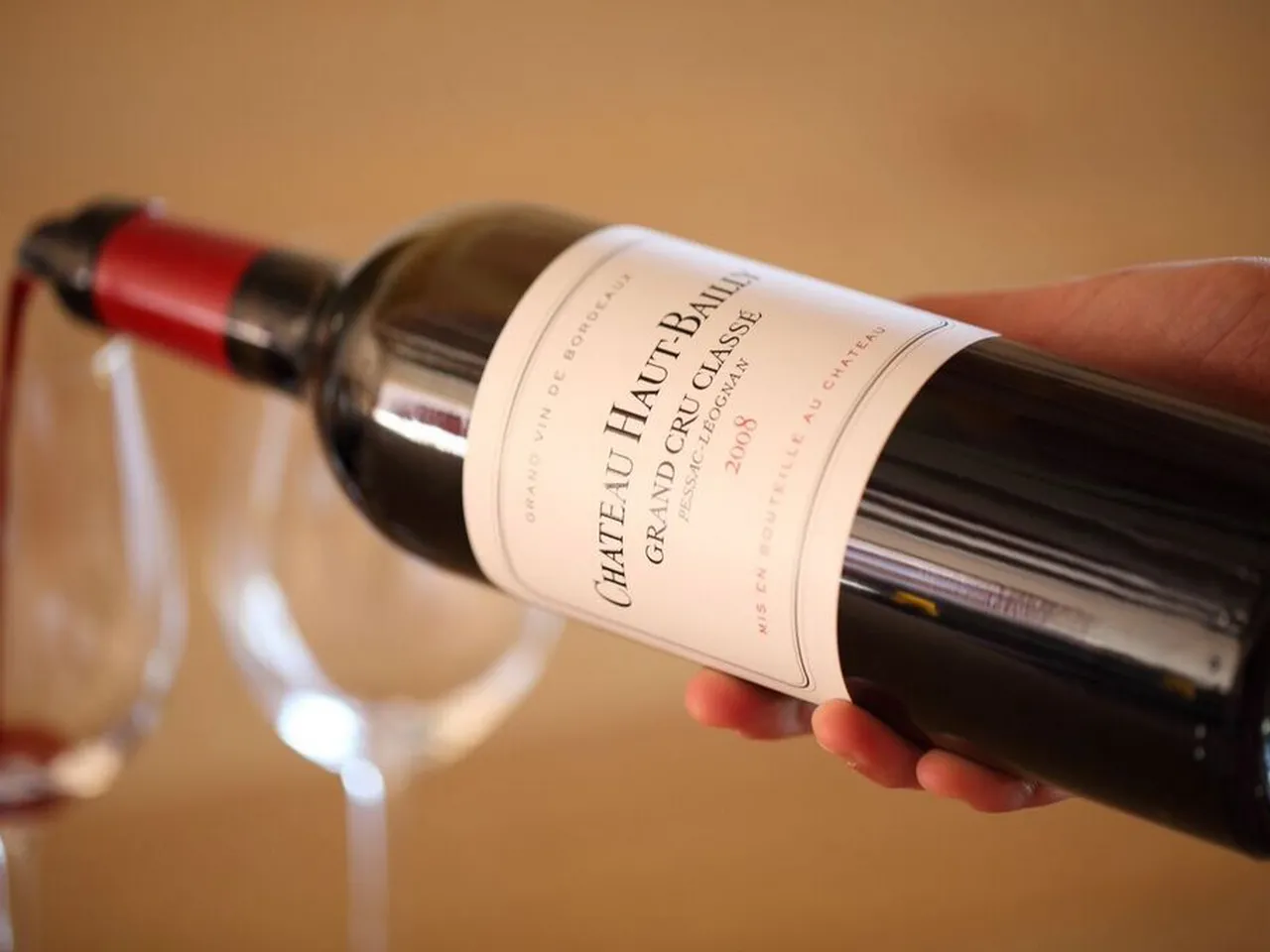 A hand holding a bottle of Château Haut-Bailly Grand Cru Classé wine from Bordeaux, with wine glasses in the background