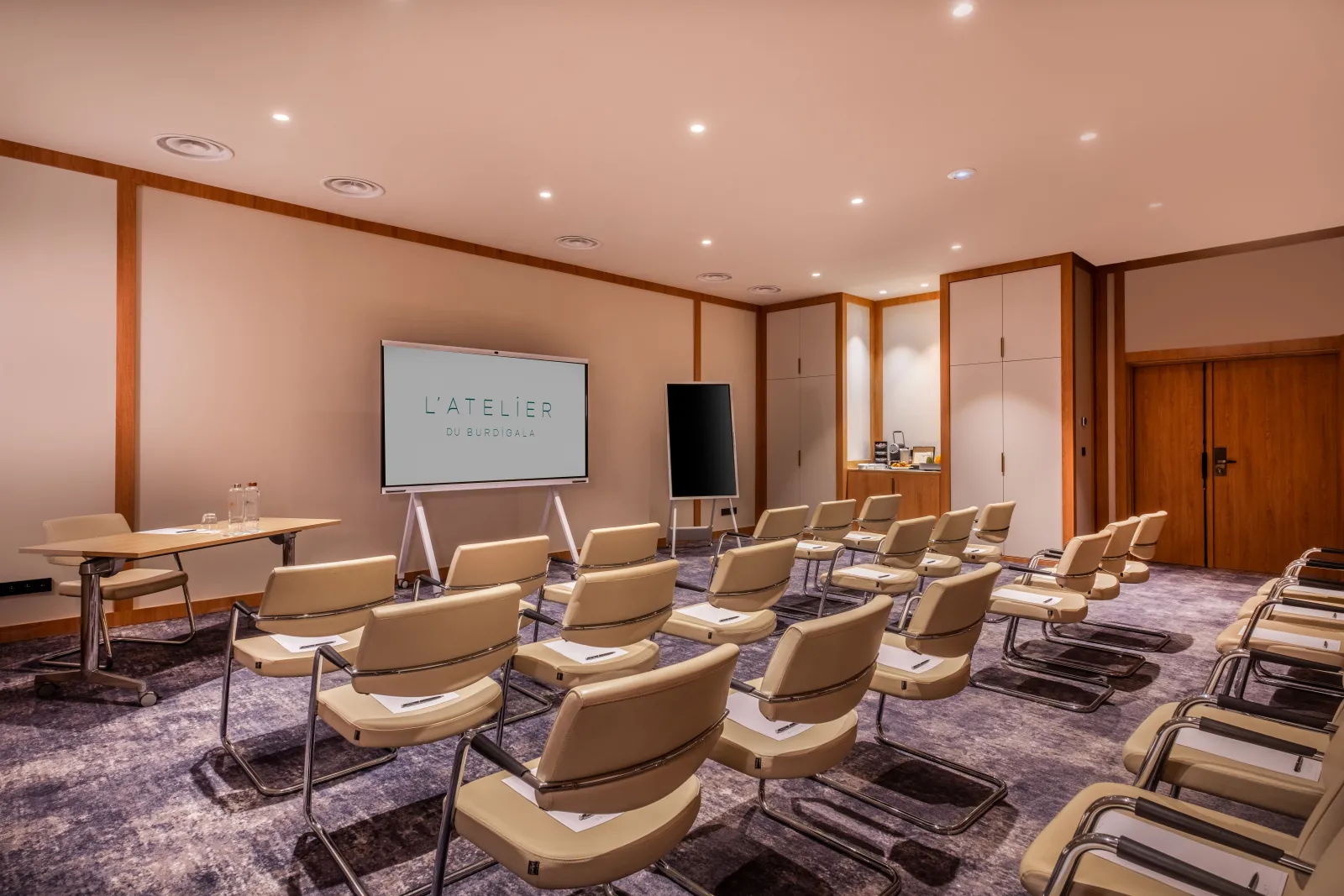 Modern conference room at Burdigala Hotel Bordeaux with beige chairs arranged in rows, projection screen displaying 'L'ATELIER DU BURDIGALA', wooden ceiling details, and warm ambient lighting