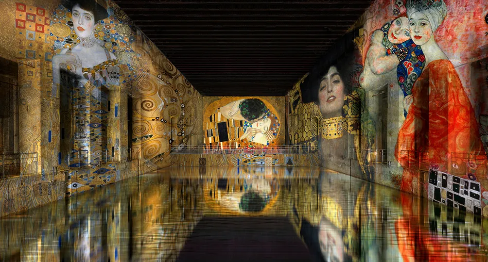Immersive digital art installation at Bassins des Lumières in Bordeaux, featuring large-scale projections of Gustav Klimt's portraits and masterpieces on warehouse walls, with reflections on water floor.