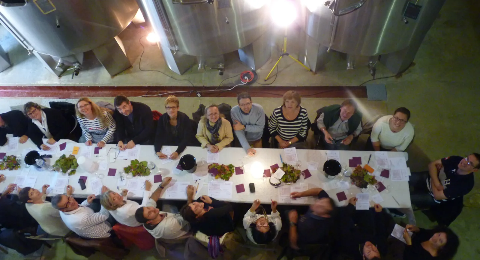 Overhead view of a wine tasting workshop at Bacchus Workshops in Bordeaux, with approximately 20 participants seated around white tables with wine glasses, tasting notes, and grape samples, under professional lighting in an industrial venue