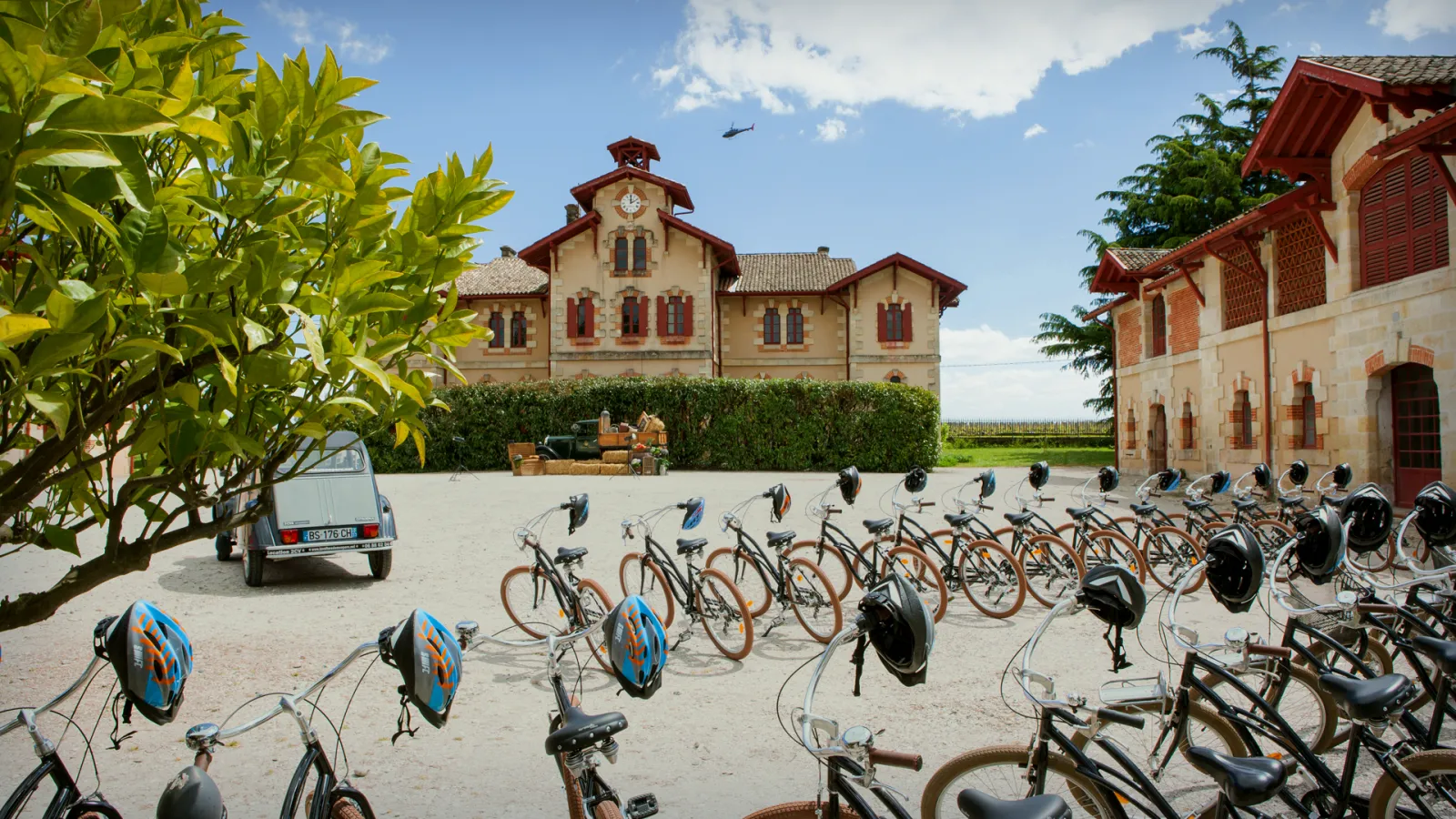 A bike rental station at a winery estate in Bordeaux, featuring multiple bicycles with helmets parked in front of traditional stone buildings with red shutters, a vintage car, and manicured gardens.