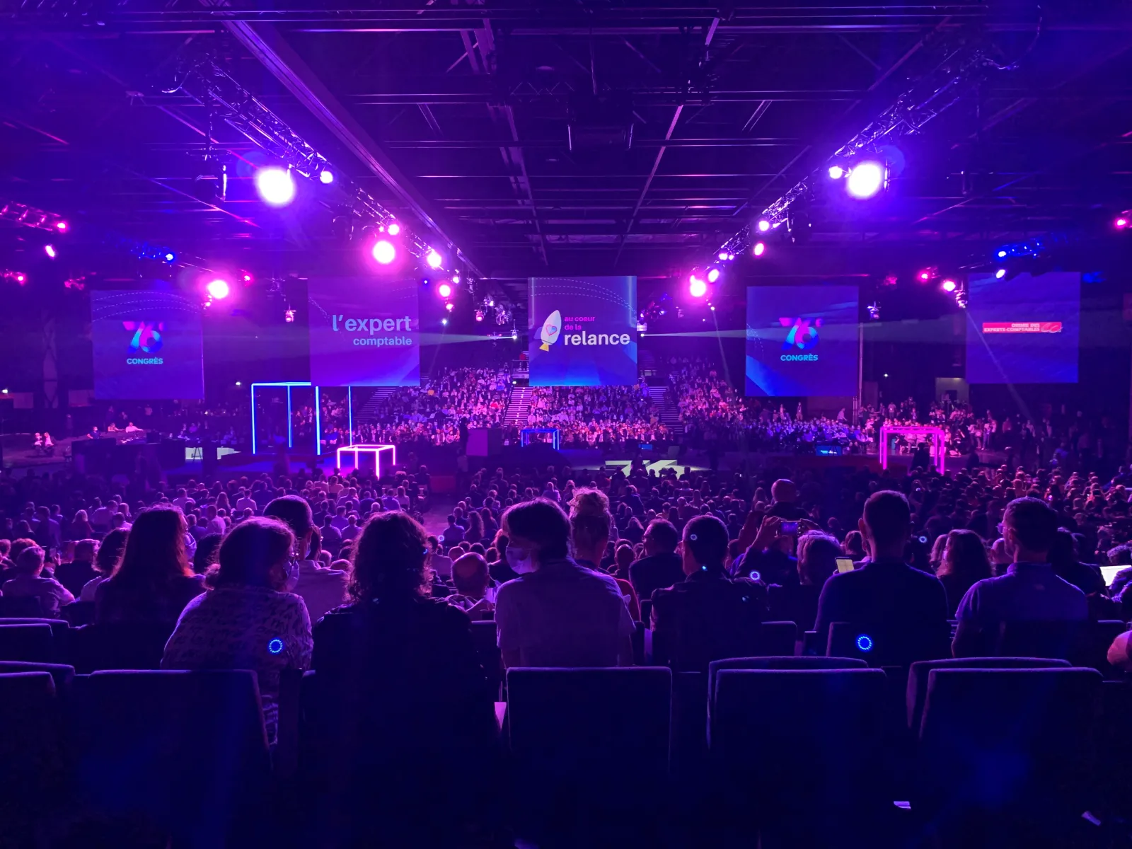 Large indoor conference or concert venue filled with hundreds of attendees seated in rows, illuminated by purple and pink stage lighting, with a main stage featuring multiple screens displaying 'reliance' branding and 'l'expert comptable' text at Palais 2