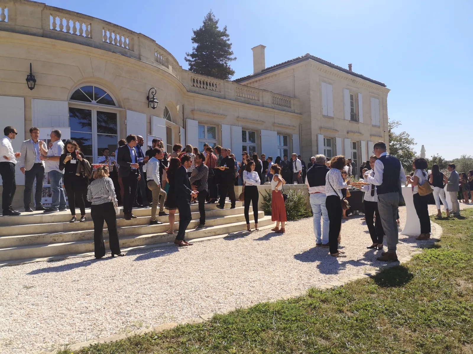 Outdoor reception event at Château Gassies in Bordeaux, with formally dressed guests gathering on a gravel courtyard in front of a classical stone mansion on a sunny day.