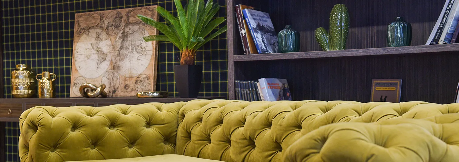 Luxury hotel interior featuring a mustard yellow tufted sofa, dark shelving with cacti and books, a vintage world map, gold decorative objects, and a geometric patterned wall backdrop.