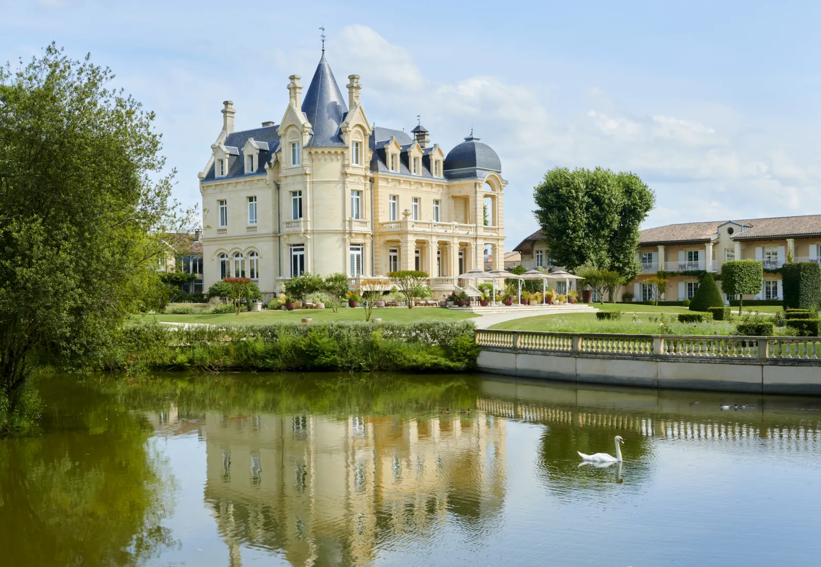 Grand Barrail castle hotel in Bordeaux, a cream-colored Victorian mansion with turrets and mansard roof reflected in a tranquil canal, surrounded by manicured gardens and greenery with a white swan on the water.
