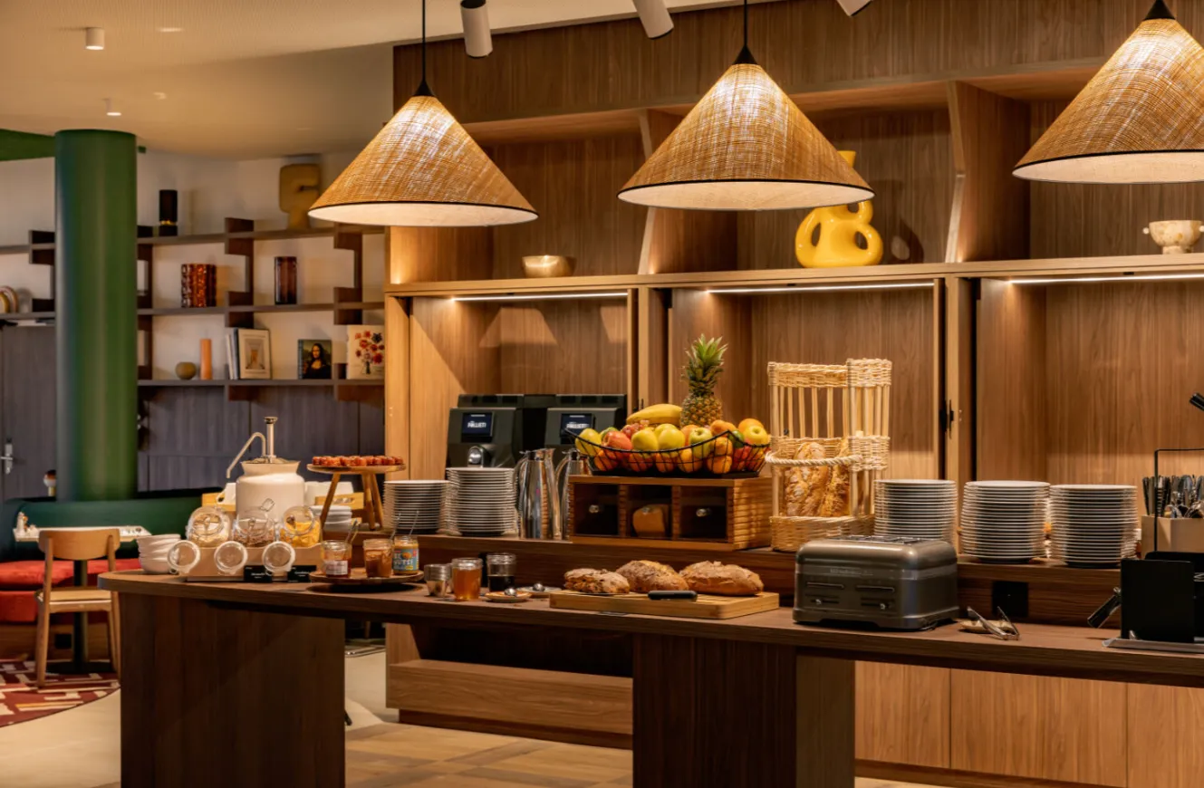Modern hotel breakfast buffet area featuring wooden pendant lights, fresh fruit display, pastries, beverages, and hospitality amenities in a warm contemporary setting.