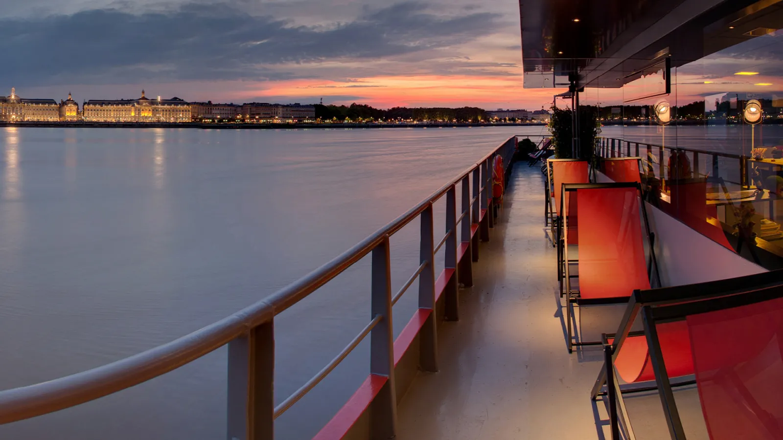 Sunset view from a Bordeaux river cruise boat deck with red chairs, overlooking the illuminated waterfront buildings and Garonne river at dusk