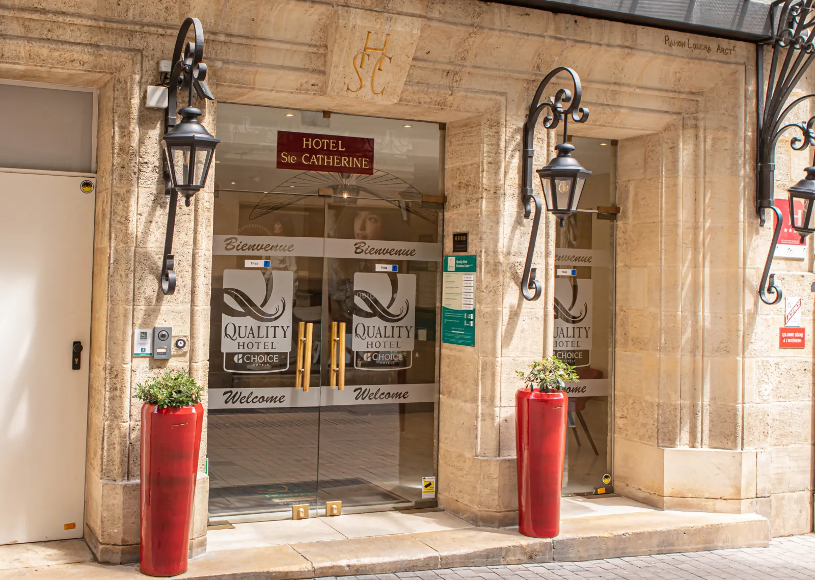 Entrance of Hotel Sainte Catherine, a Quality Hotel in Bordeaux city center, featuring glass doors with 'Welcome' and 'Bienvenue' text, decorative wrought iron lanterns, and red planters with greenery on the stone facade.