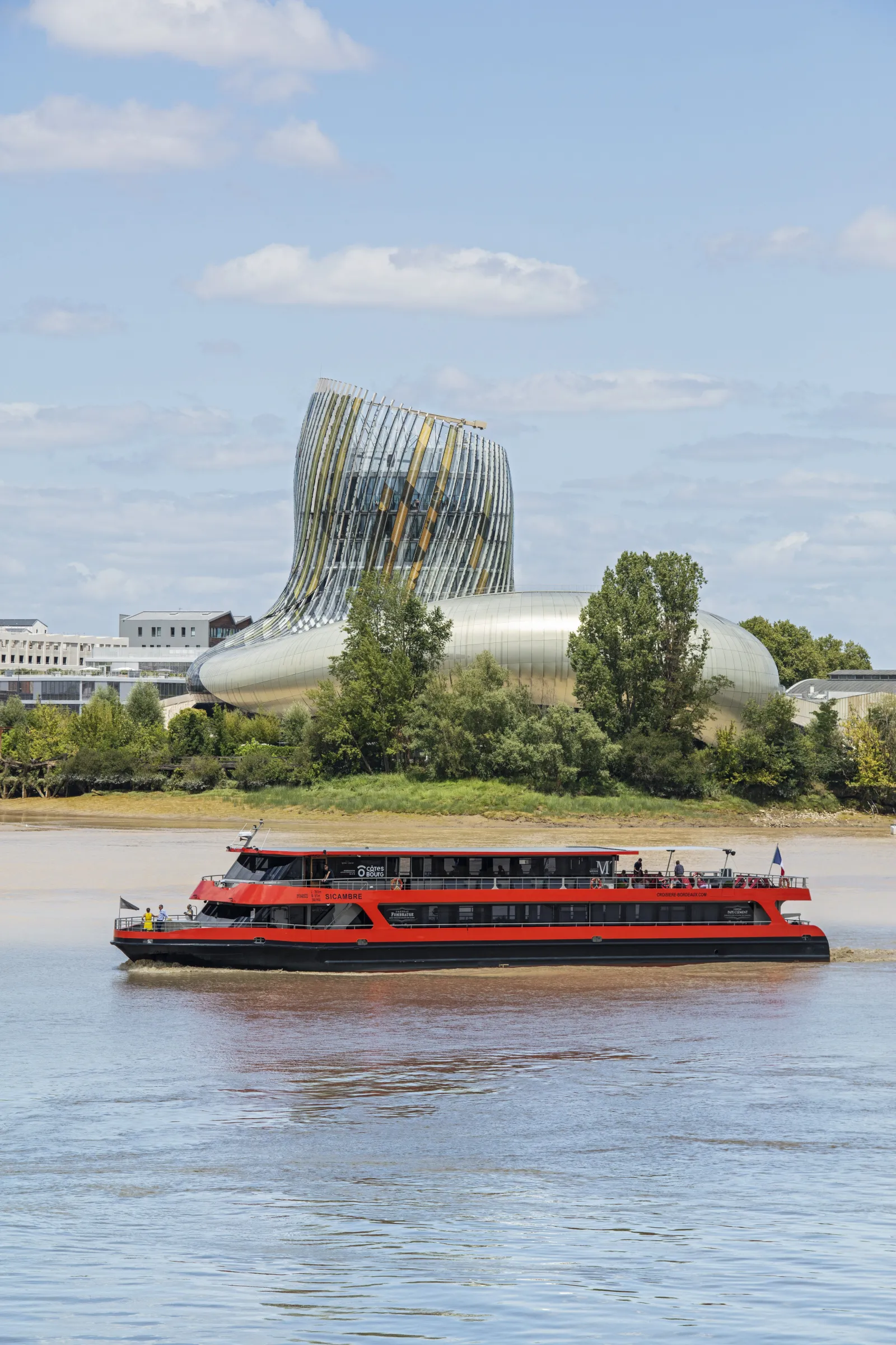 A red and black river cruise boat traveling on the Garonne River in Bordeaux, with the modern La Cité du Vin wine museum building featuring a distinctive tilted glass and metal structure in the background.