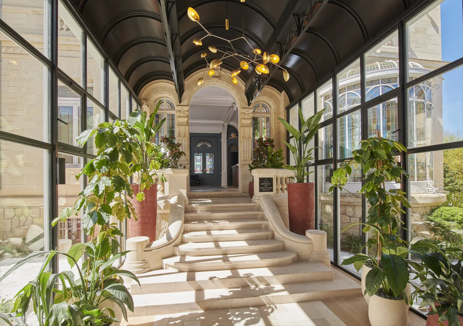 Elegant covered passageway with glass panels and black metal framework leading to the entrance of Château Hôtel Grand Barrail in Bordeaux, adorned with climbing plants in terracotta pots and golden decorative lighting fixtures.