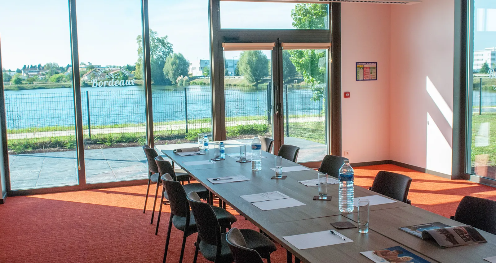 Modern conference room with large windows overlooking a river and garden, featuring a long table with black chairs, water bottles, and documents, with red carpeting and pink walls.