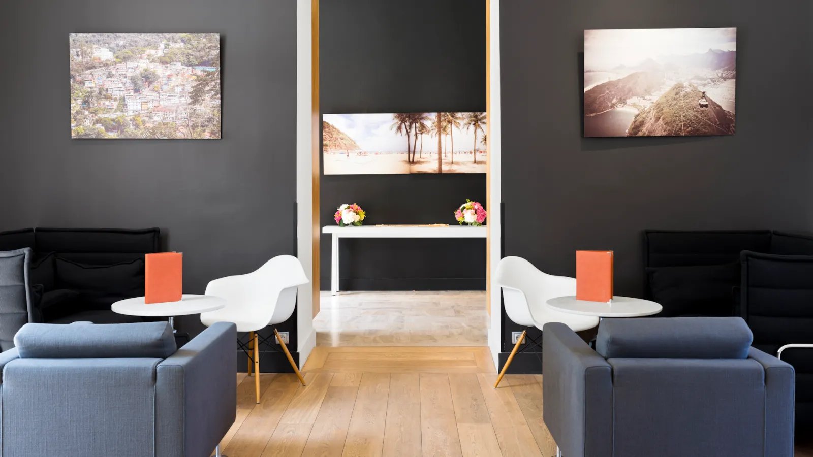 Modern hotel lounge at Le Saint-James Bordeaux featuring symmetrical seating areas with gray armchairs, white round tables with orange accents, dark walls with landscape photography, a central display area with floral arrangements, wooden flooring, and co