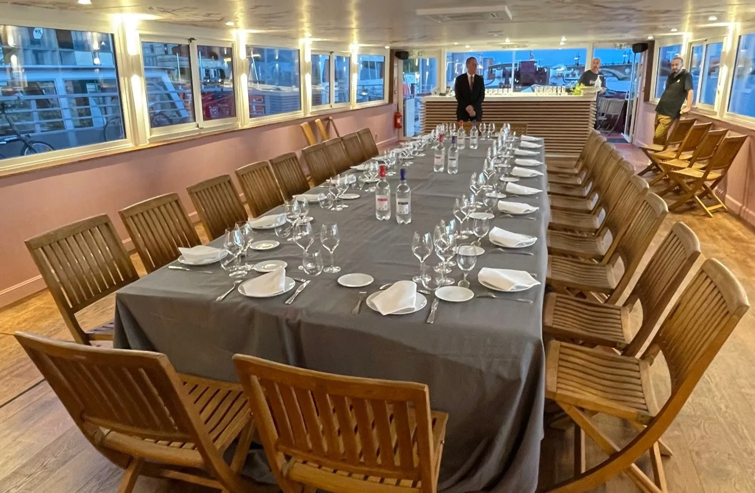Elegant dining room on a yacht with long table set for formal dinner, wooden chairs, place settings, glasses, and panoramic windows overlooking a harbor at dusk
