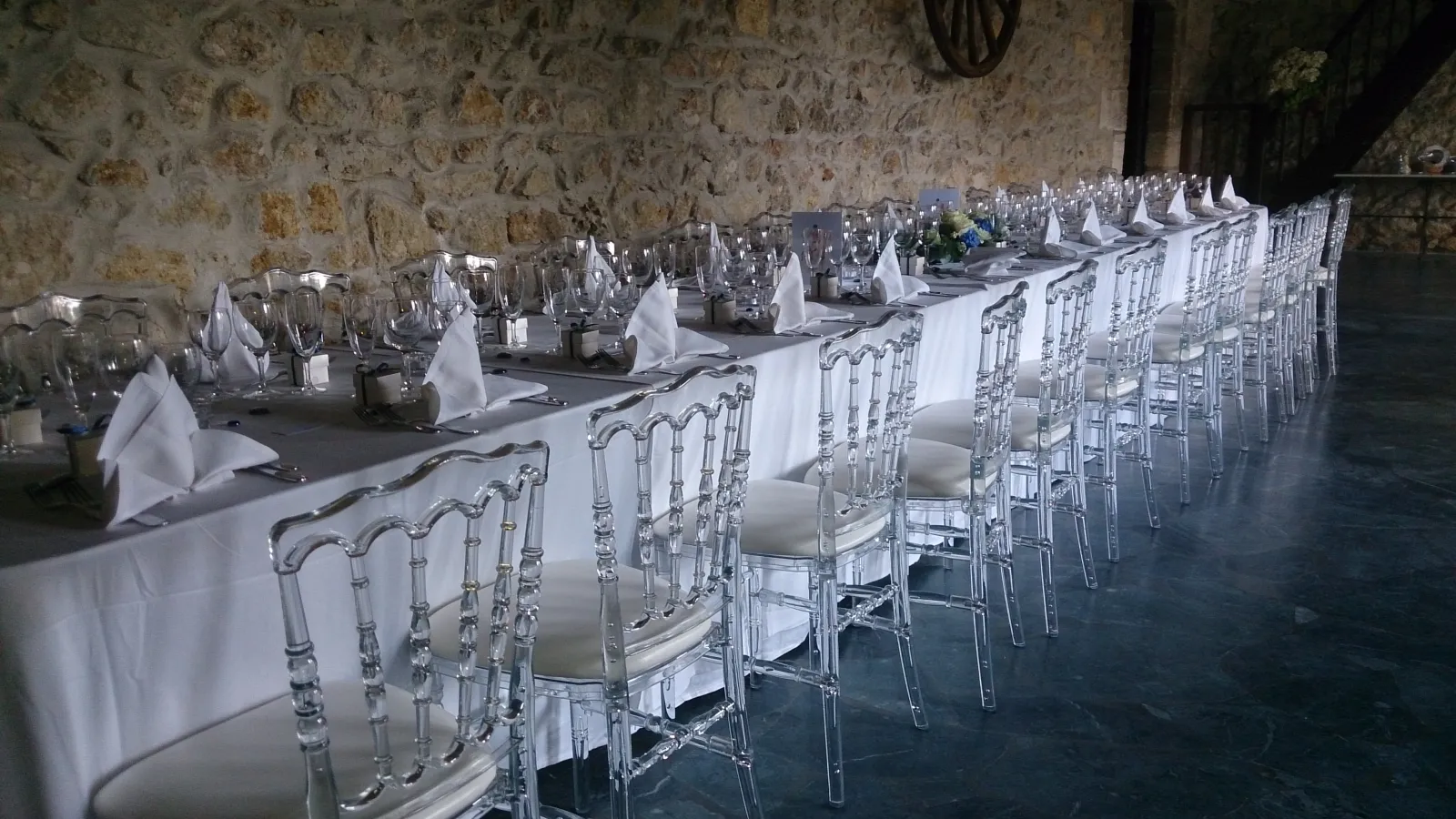 Long elegant banquet table with white tablecloths, transparent chiavari chairs with white cushions, folded white napkins, and place settings in a stone-walled venue prepared for a formal event