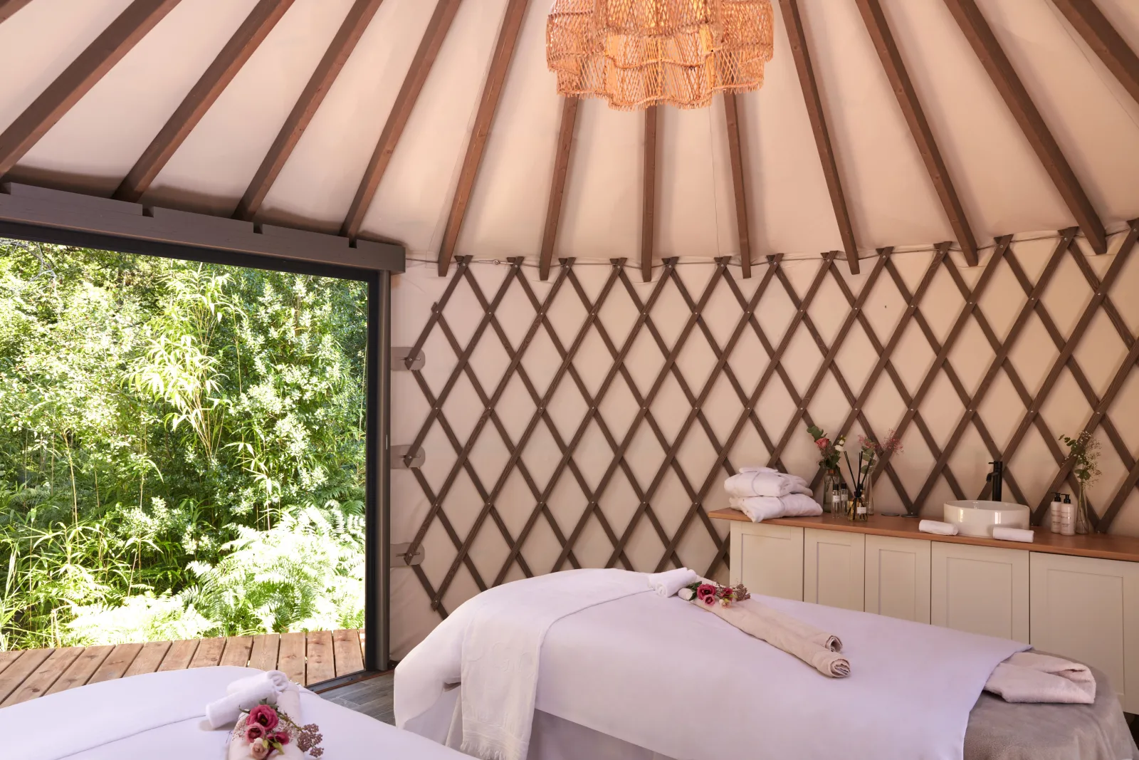 Interior of a luxury spa yurt with two massage tables, cream-colored fabric walls with wooden lattice details, a wooden ceiling with beams, and an open door overlooking a lush green garden.