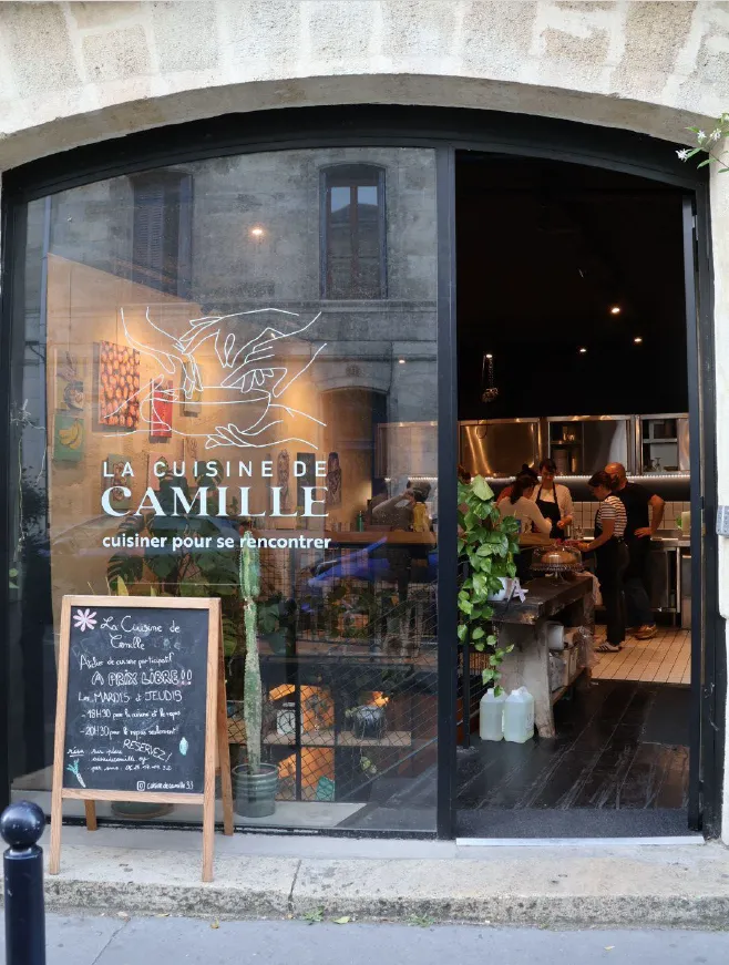 Storefront of La Cuisine de Camille restaurant in Bordeaux, featuring large curved windows with illuminated neon logo, diners visible inside, and a wooden A-frame sign on the sidewalk.
