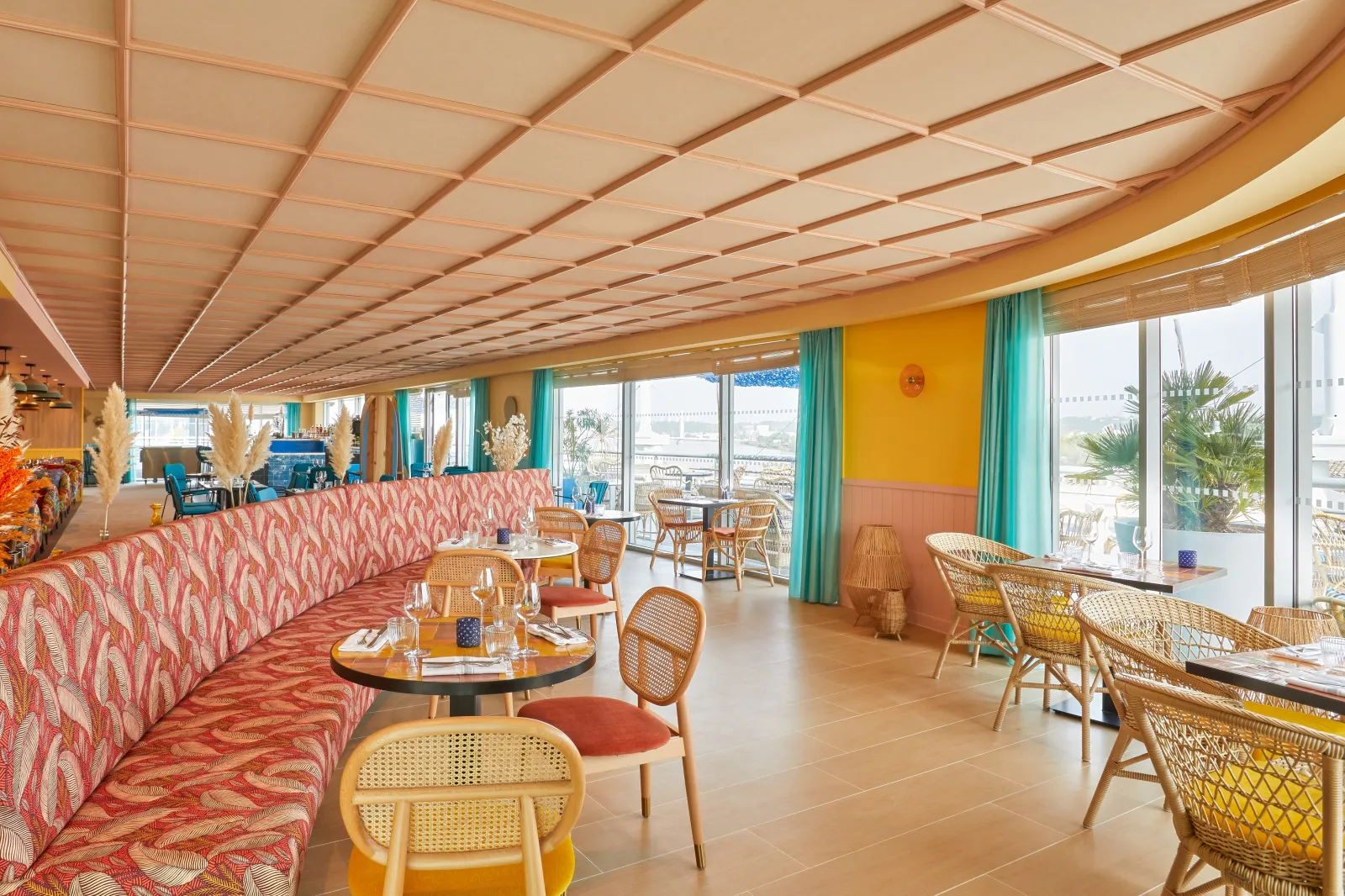 Modern hotel lounge and dining area with colorful furnishings, floor-to-ceiling windows overlooking Bordeaux, coral-patterned seating, wicker chairs, and a geometric ceiling design with warm lighting.
