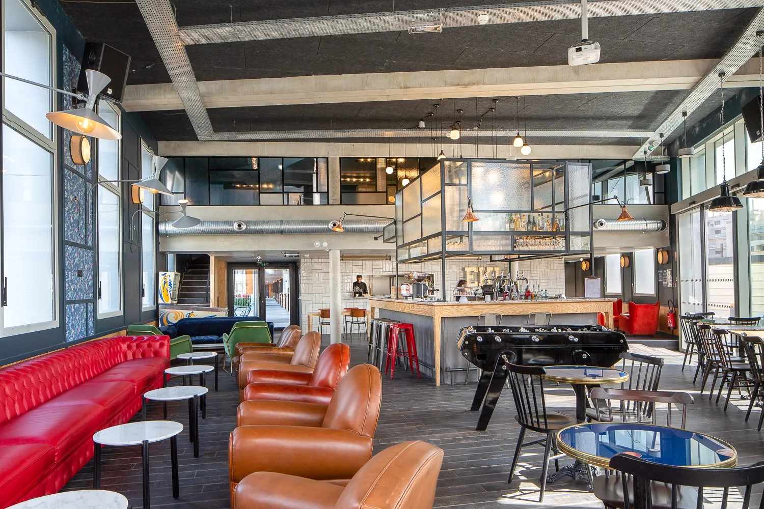 Modern lounge and recreation area at Eklo Bordeaux hotel featuring red and cognac leather seating, foosball table, bar counter, and industrial ceiling with exposed beams