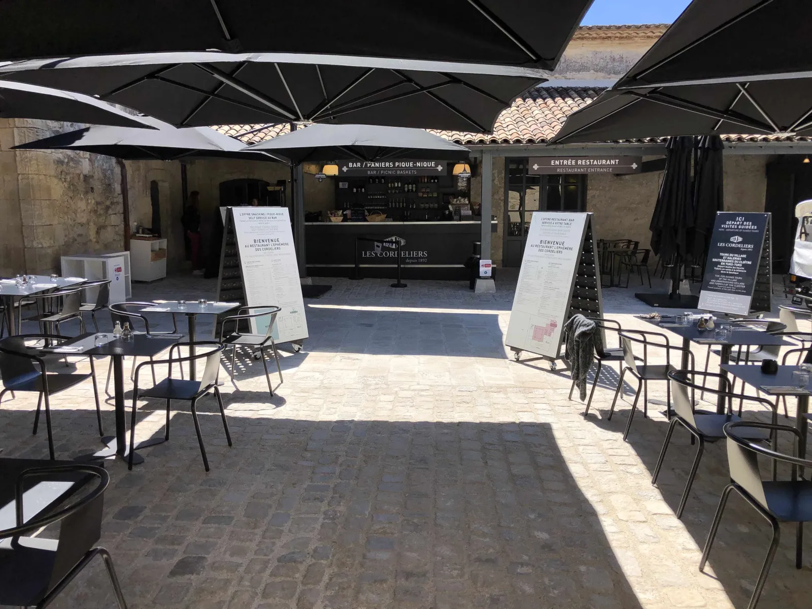 Outdoor terrace of Les Cordeliers restaurant in Bordeaux, featuring black umbrellas, modern metal chairs, stone paving, and a bar entrance with welcome signs.
