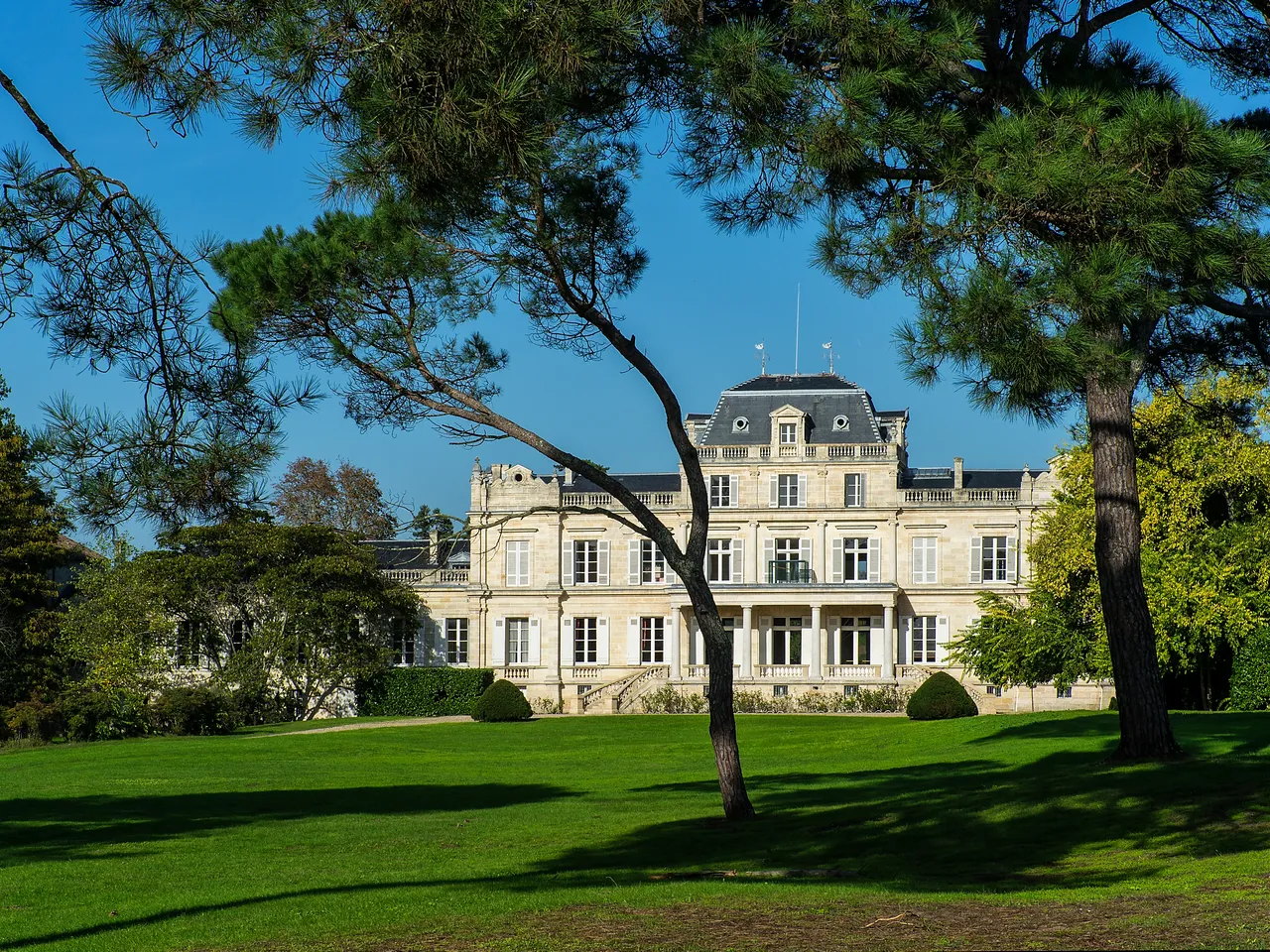 Château Giscours, a grand 19th-century mansion with cream-colored stone facade, mansard roof, and manicured grounds framed by mature trees under clear blue sky in Bordeaux.