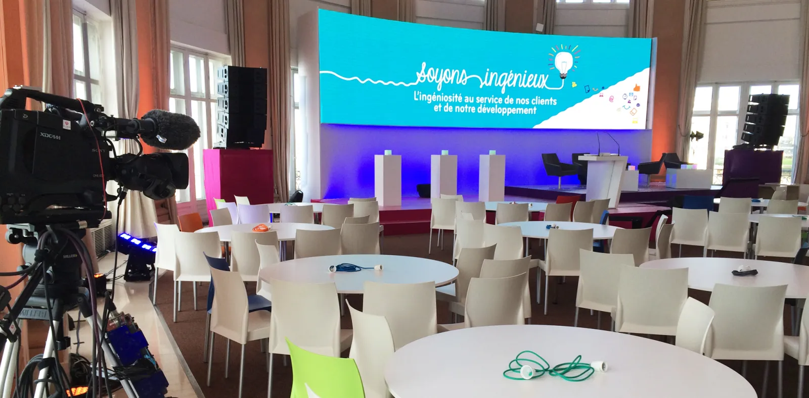 Conference room setup in Bordeaux with white chairs around round tables, a large turquoise and purple display screen showing 'Soyons ingénieux' branding, professional video camera on the left, and windows with orange-tinted curtains in the background.