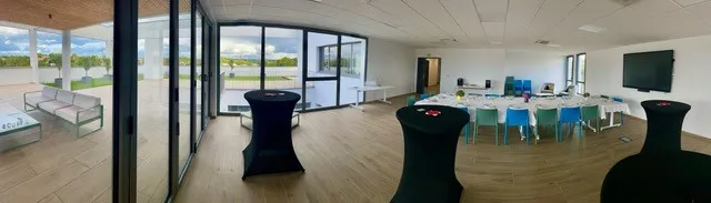Modern event space with wooden flooring, floor-to-ceiling windows overlooking countryside, black high-top tables, and a bright workspace area with turquoise seating in the background.