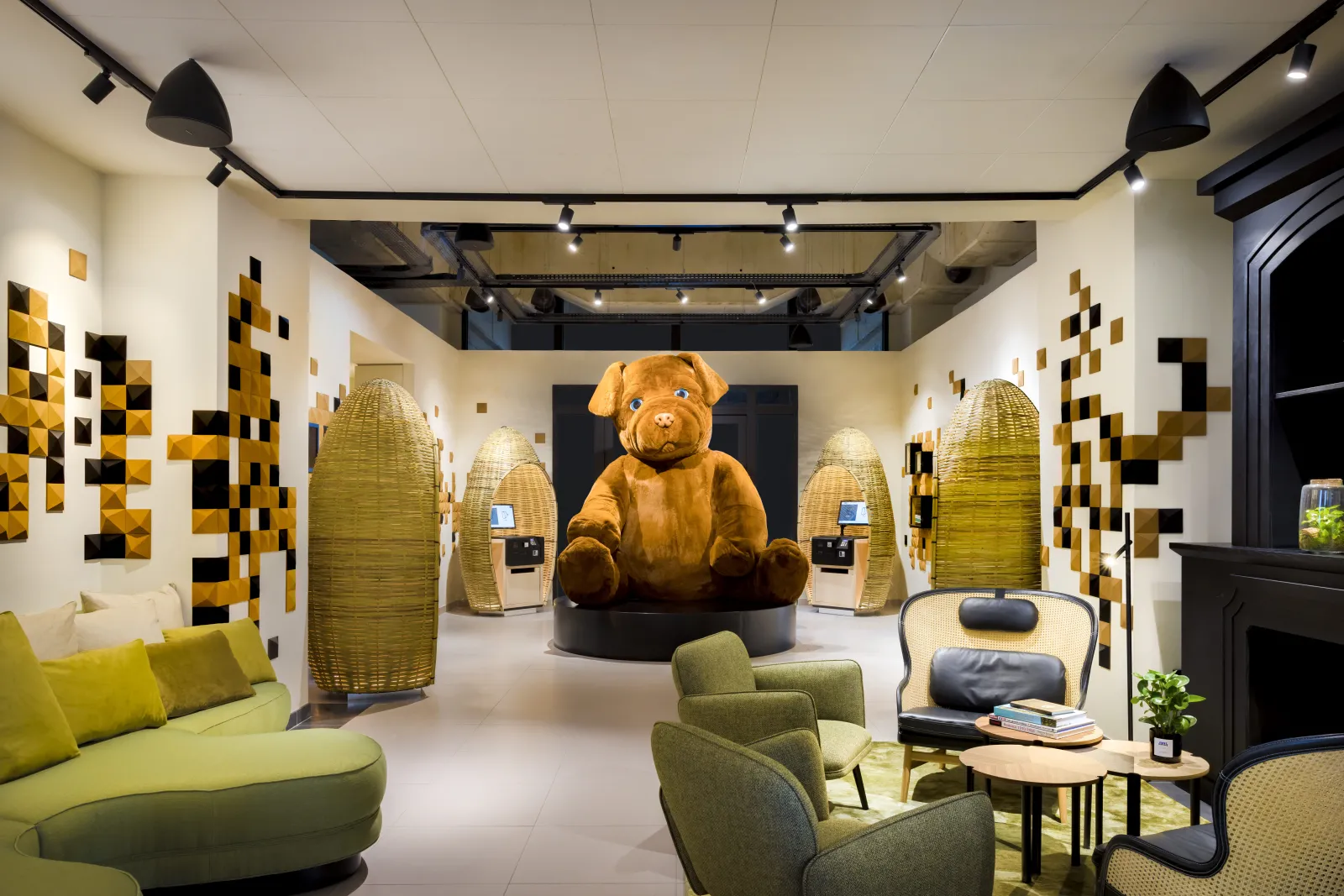 Modern hotel lobby lounge featuring a large golden bear mascot in the center, surrounded by yellow and black geometric wall patterns, contemporary seating with yellow and green upholstered chairs, woven basket-shaped accent elements, and track lighting th