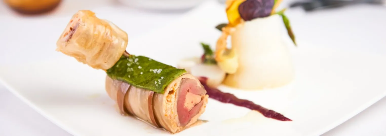 Elegant gourmet appetizer skewers featuring foie gras wrapped in bacon with green leaf garnish, served on a white plate with decorative elements